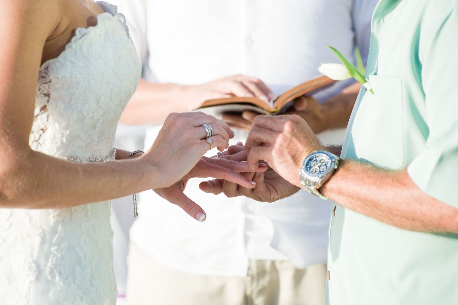 Couple exchanging wedding rings during a beach ceremony, with an officiant holding a book in the background.