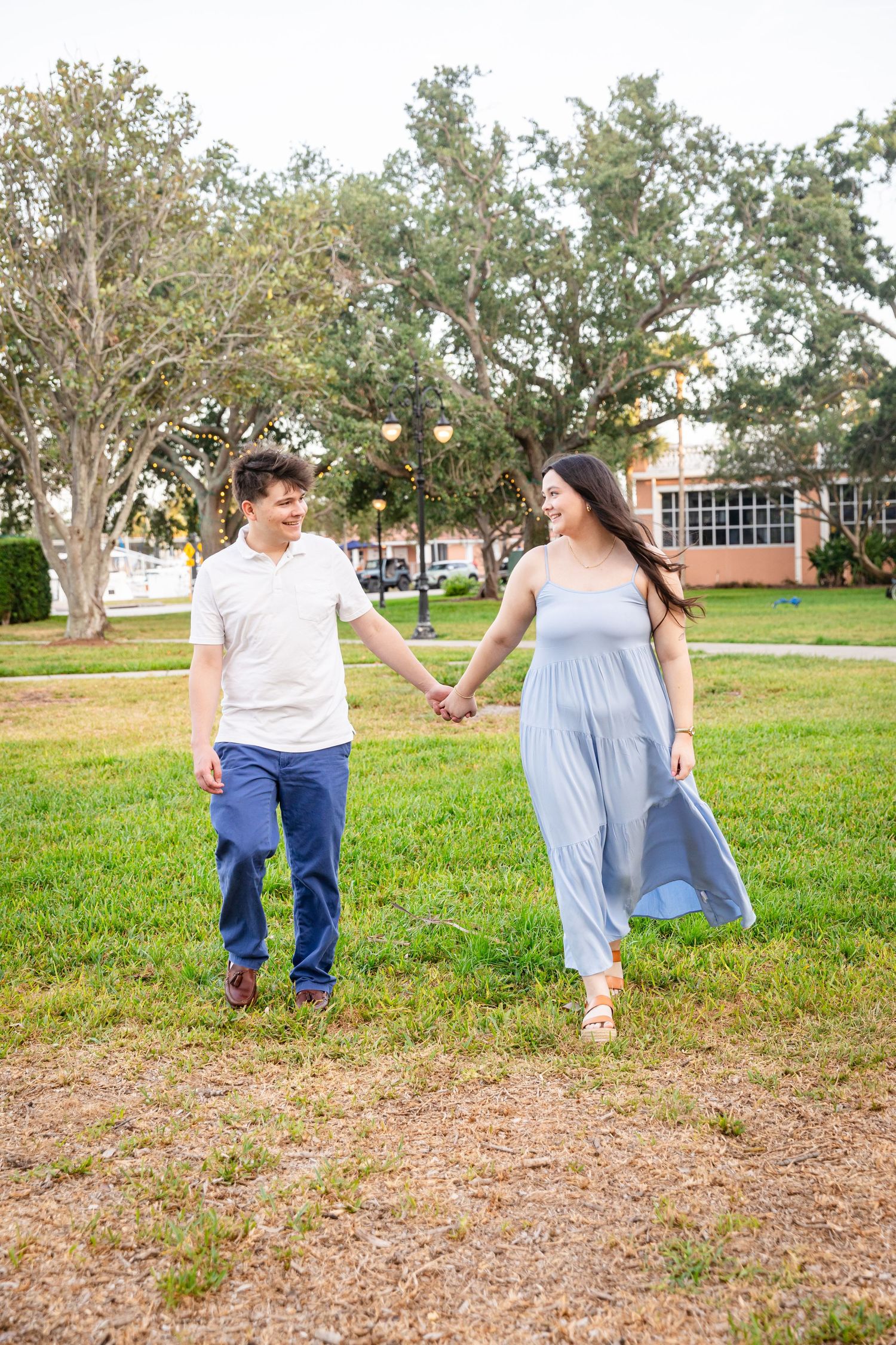 A smiling couple holds hands while walking through a lush green park, man in white polo and blue pants, woman in light blue maxi dress.