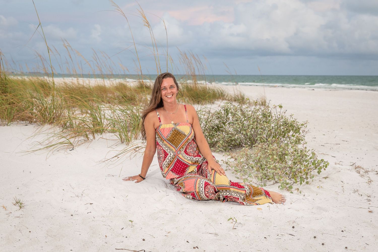 A smiling woman in a colorful maxi dress sits on white sand beach with sea oats and ocean in the background.