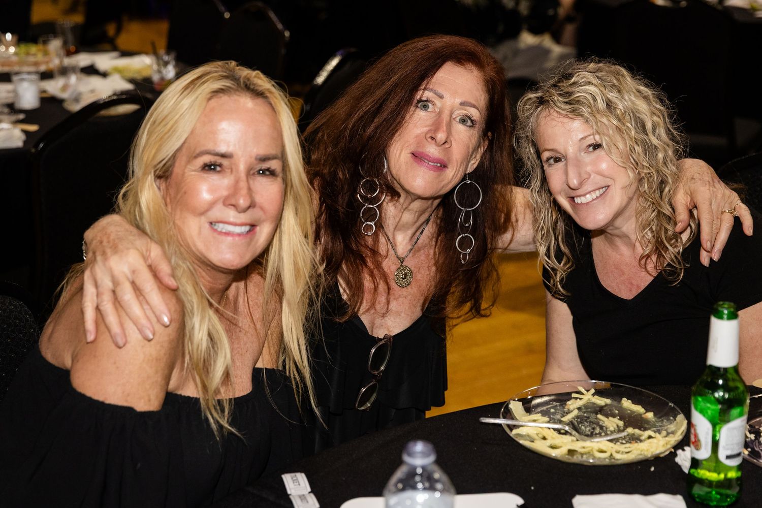 Three smiling blonde women in black outfits pose together at a dinner table at a social event.