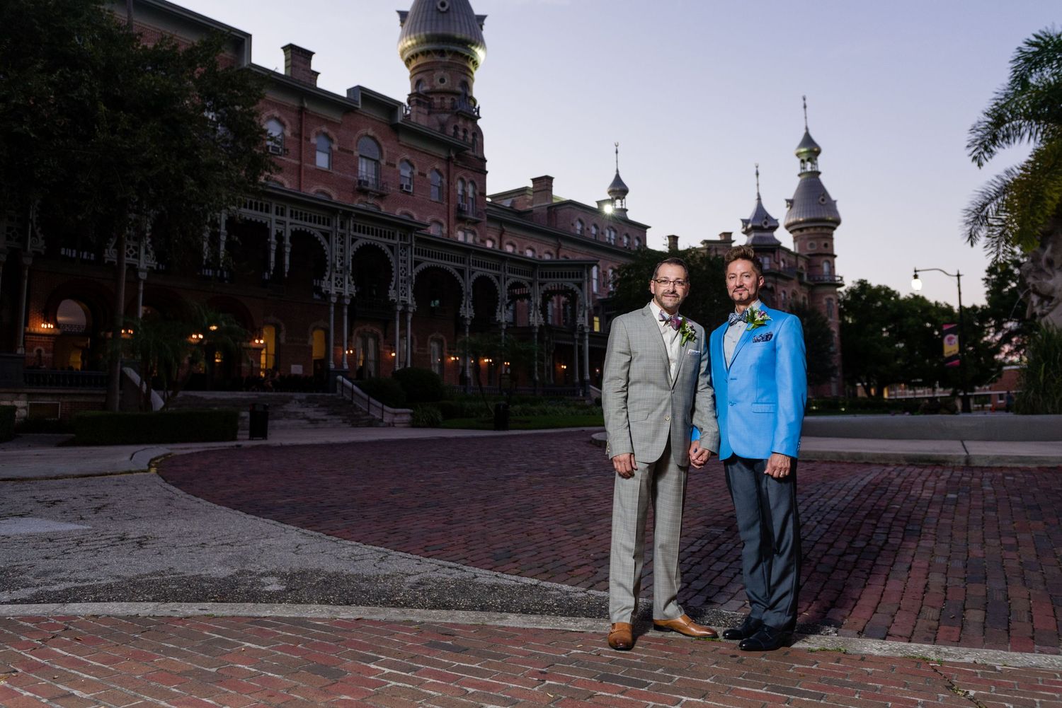 Two men in suits pose at dusk outside a historic brick building with ornate architecture and towers.