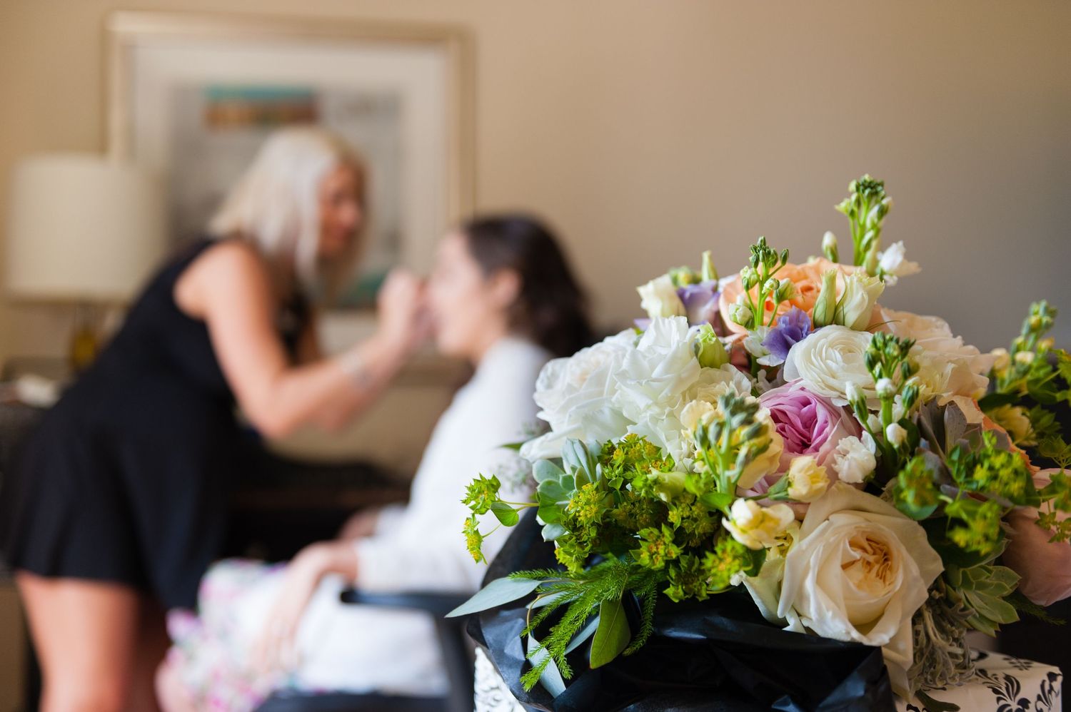 Bridal bouquet of white and pastel roses in foreground with two women preparing for a wedding in background.