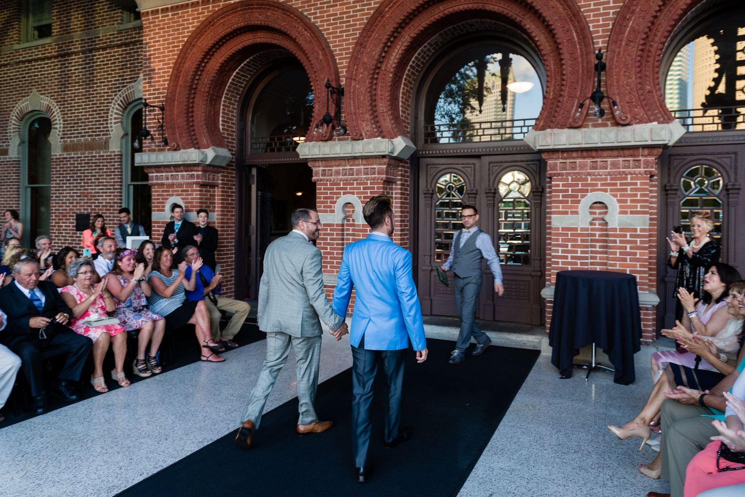 Two men in blue suits walk down a black runway at an outdoor fashion show beside a brick building.