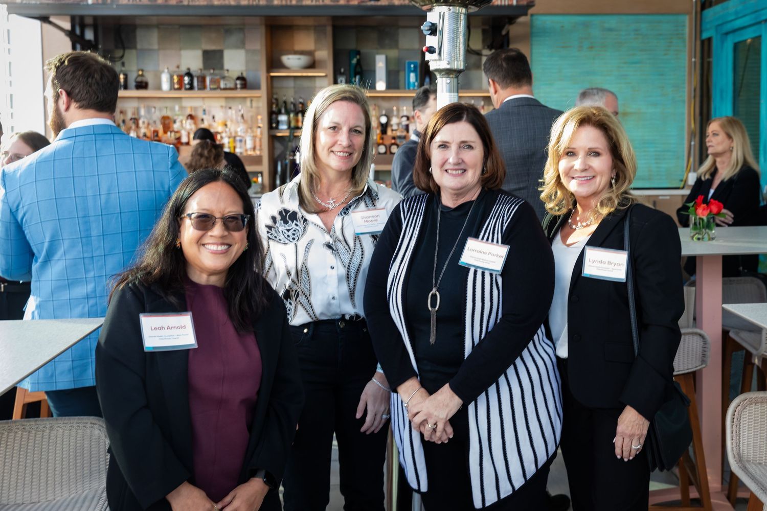 Four professional women smiling together at a networking event, wearing name badges in a modern restaurant venue.
