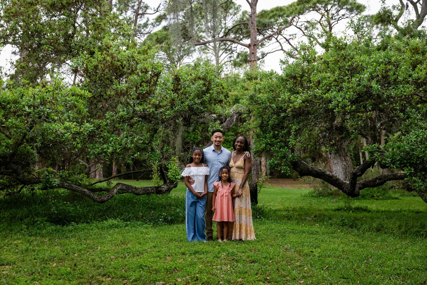 Family of four poses together on lush green grass surrounded by sprawling oak trees in a scenic outdoor setting.
