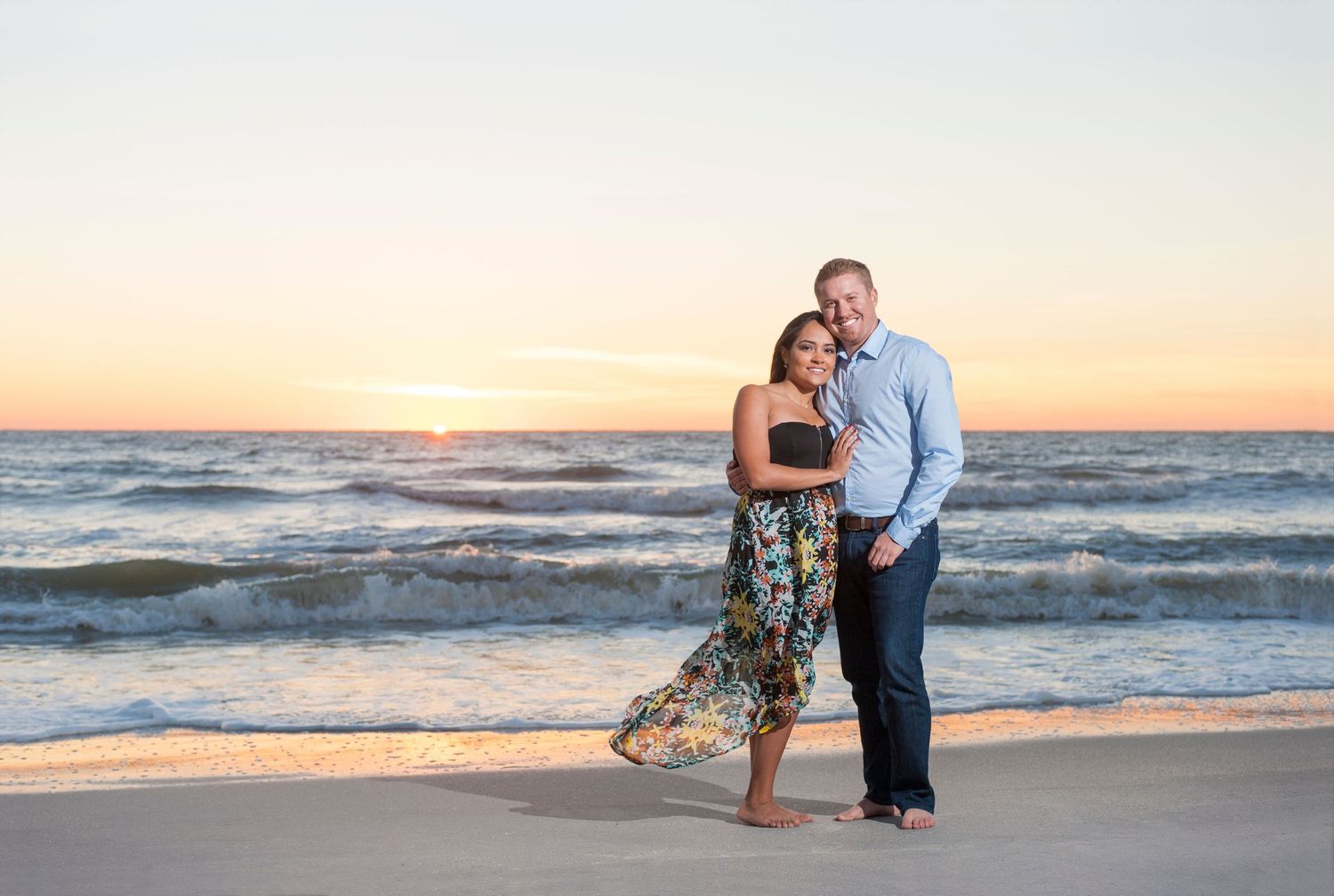 Couple embracing on a sandy beach at sunset with ocean waves and colorful sky in the background.