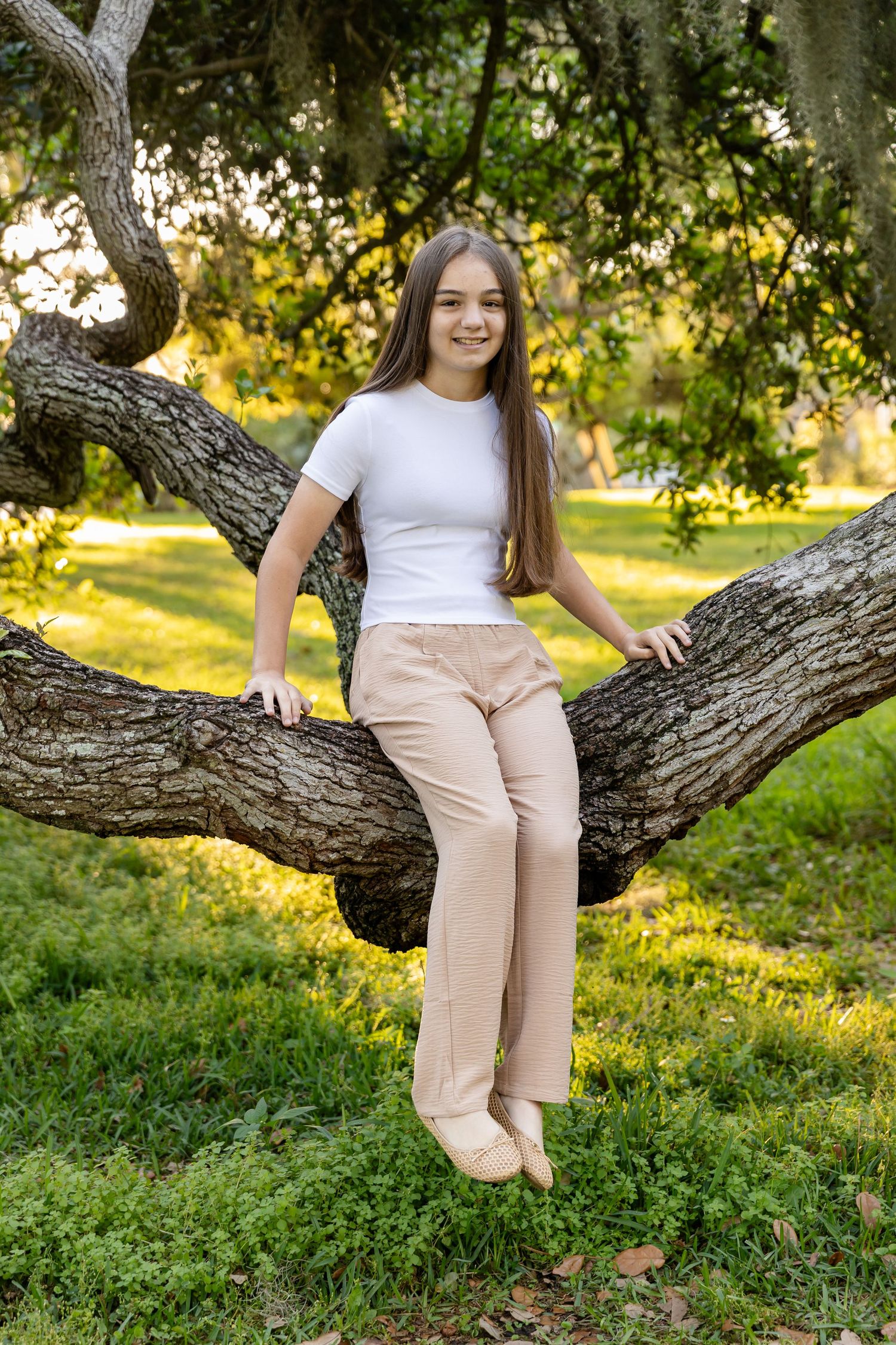 A smiling teenage girl with long brown hair sits on a curved tree branch in a lush green park.