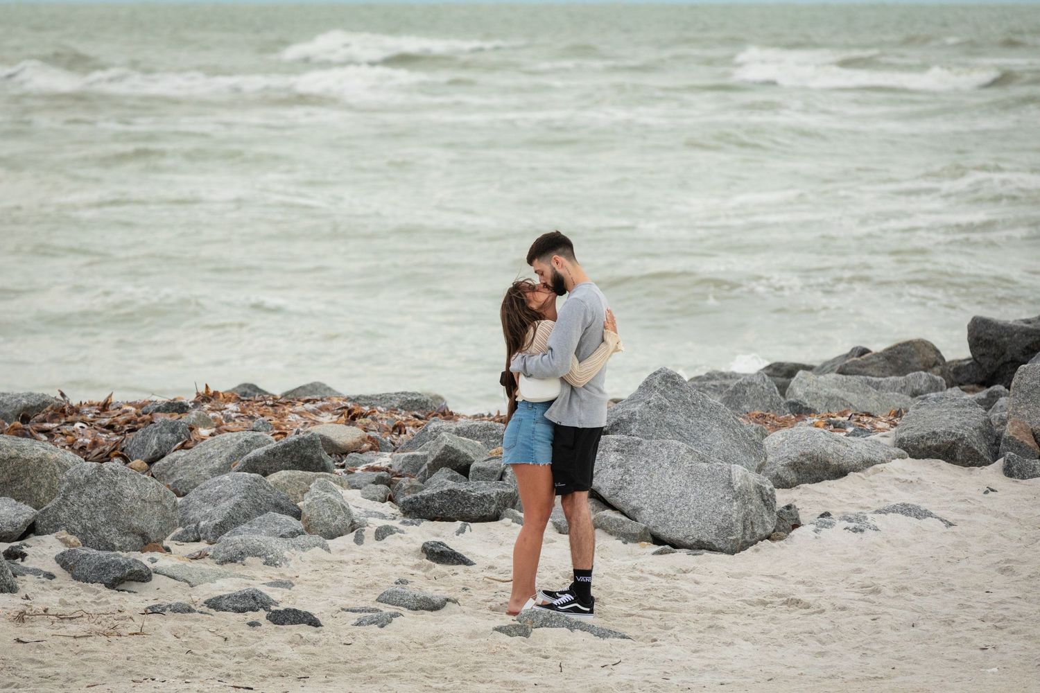 Romantic couple embracing on a rocky beach shore with ocean waves in the background.