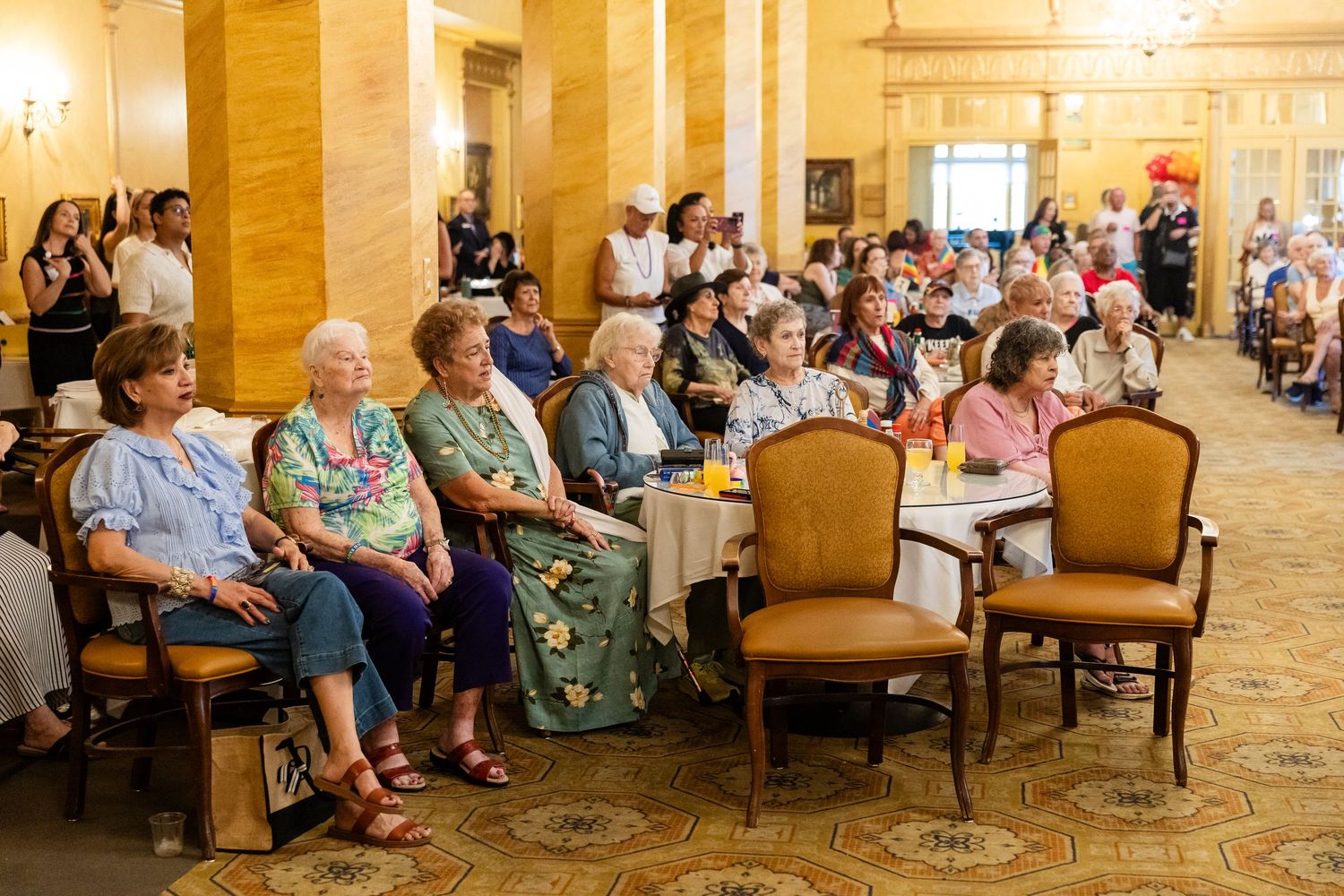 Elderly audience members seated at tables in ornate ballroom watching a live performance together.