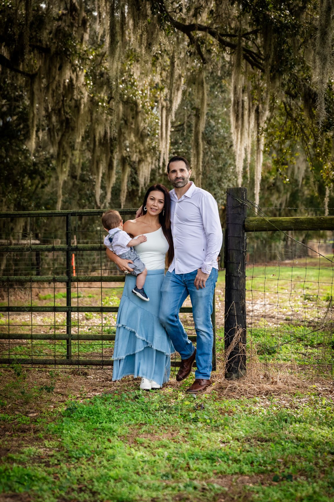 A happy family with a baby poses by a rustic farm gate under Spanish moss-draped trees.