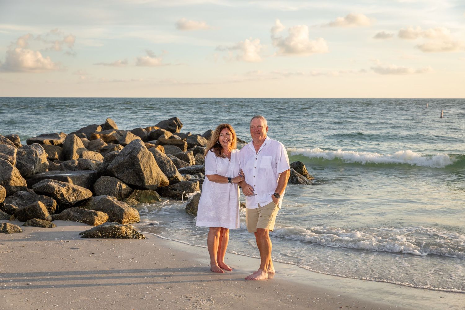 A happy couple in white outfits poses on a sandy beach near rocky jetty with ocean waves at sunset.
