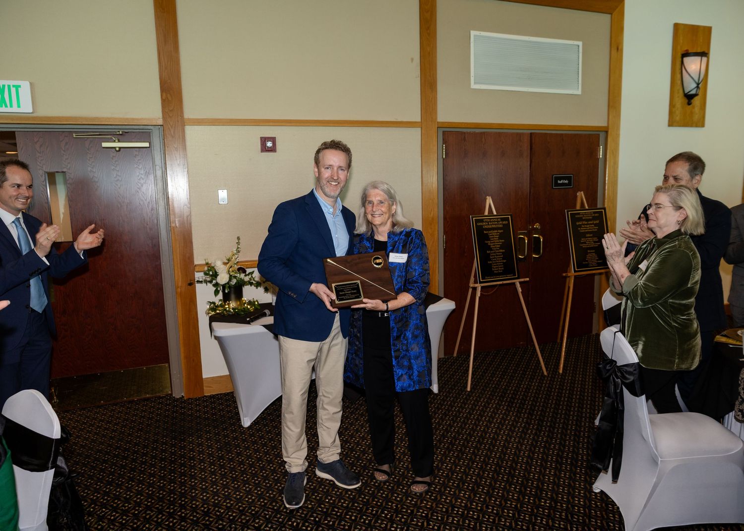 Man and woman posing together holding a wooden plaque award at a formal recognition ceremony indoors.