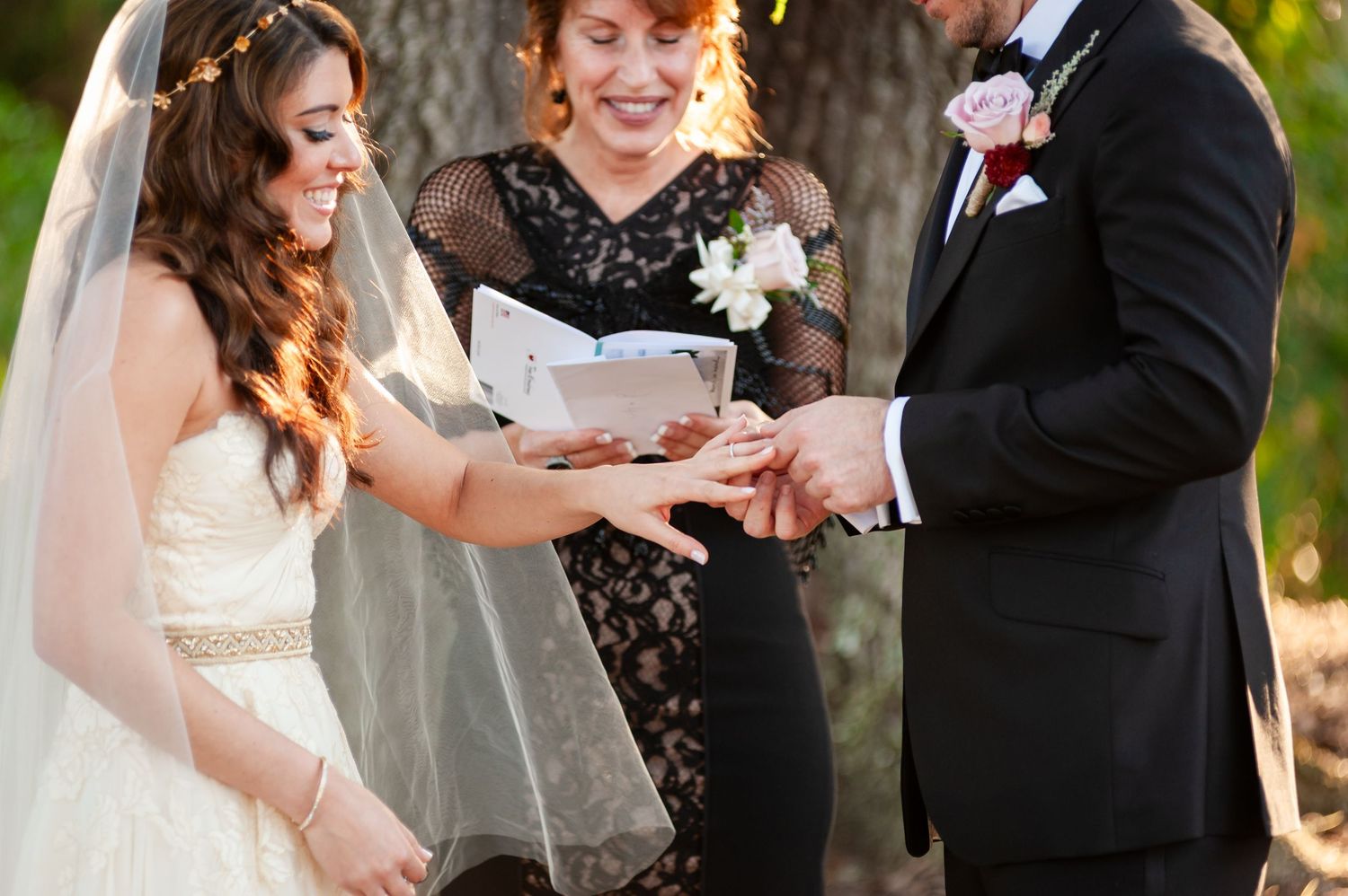 Groom places wedding ring on bride's finger as smiling officiant reads from ceremony book outdoors.