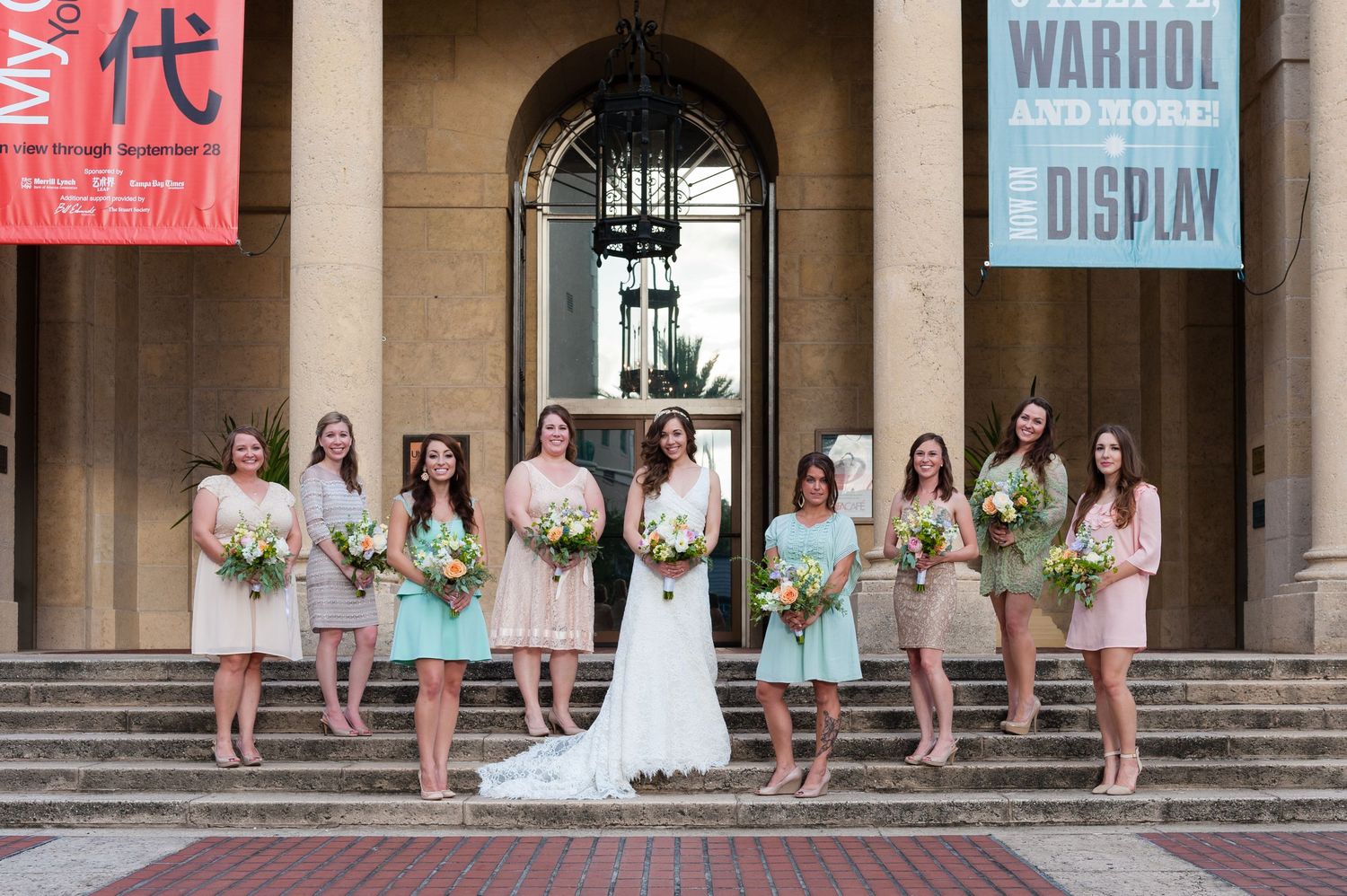 Bride and bridesmaids in mismatched pastel dresses pose on steps outside a museum with Warhol exhibit banners Museum of Fine Arts, St. Pete.