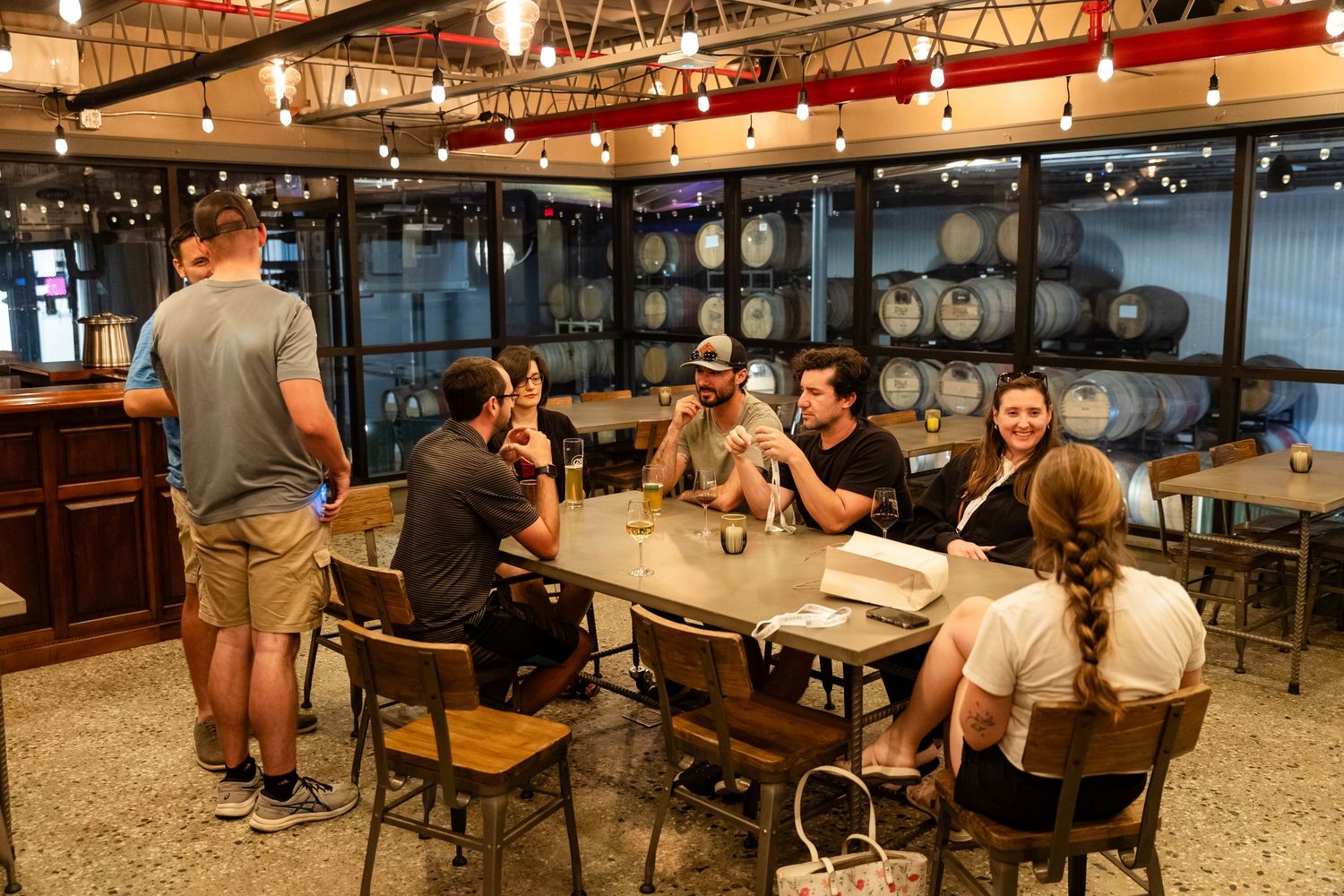 Group of friends enjoying drinks at a rustic brewery taproom with wooden barrels and warm Edison bulb lighting.