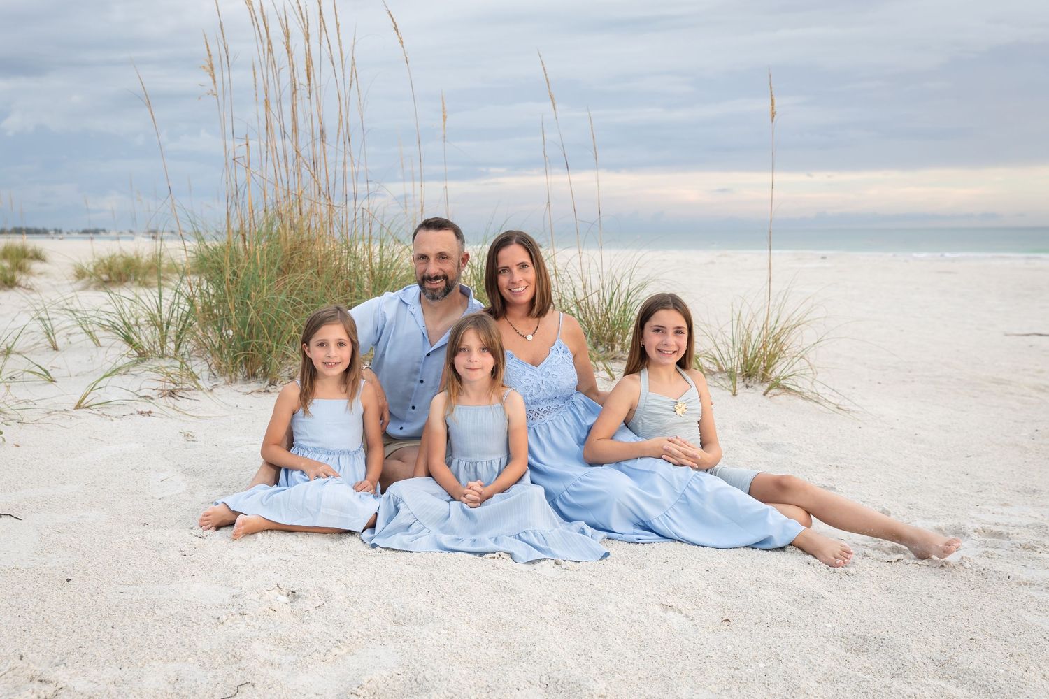 Family of five posing on white sand beach, parents with three daughters in matching light blue dresses, sea oats in background.