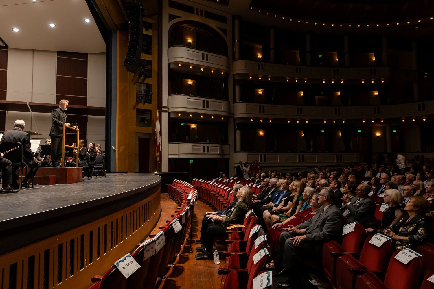 Orchestra conductor performing on stage before large audience in elegant theater venue.