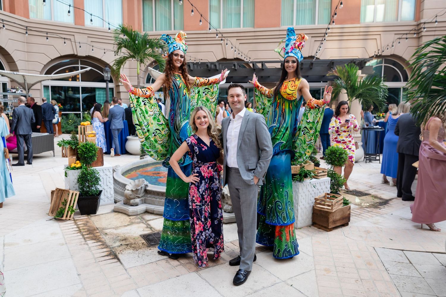 Couple poses with stilt walkers in vibrant mermaid costumes at an elegant outdoor gala event.