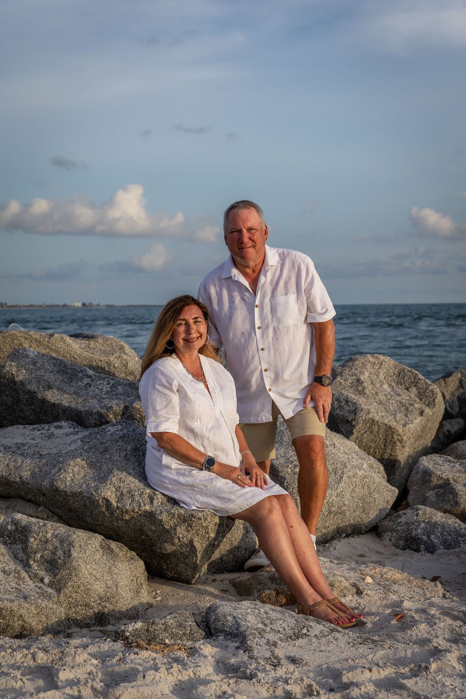 Smiling couple in white linen outfits posing on rocky Florida beach at golden hour.