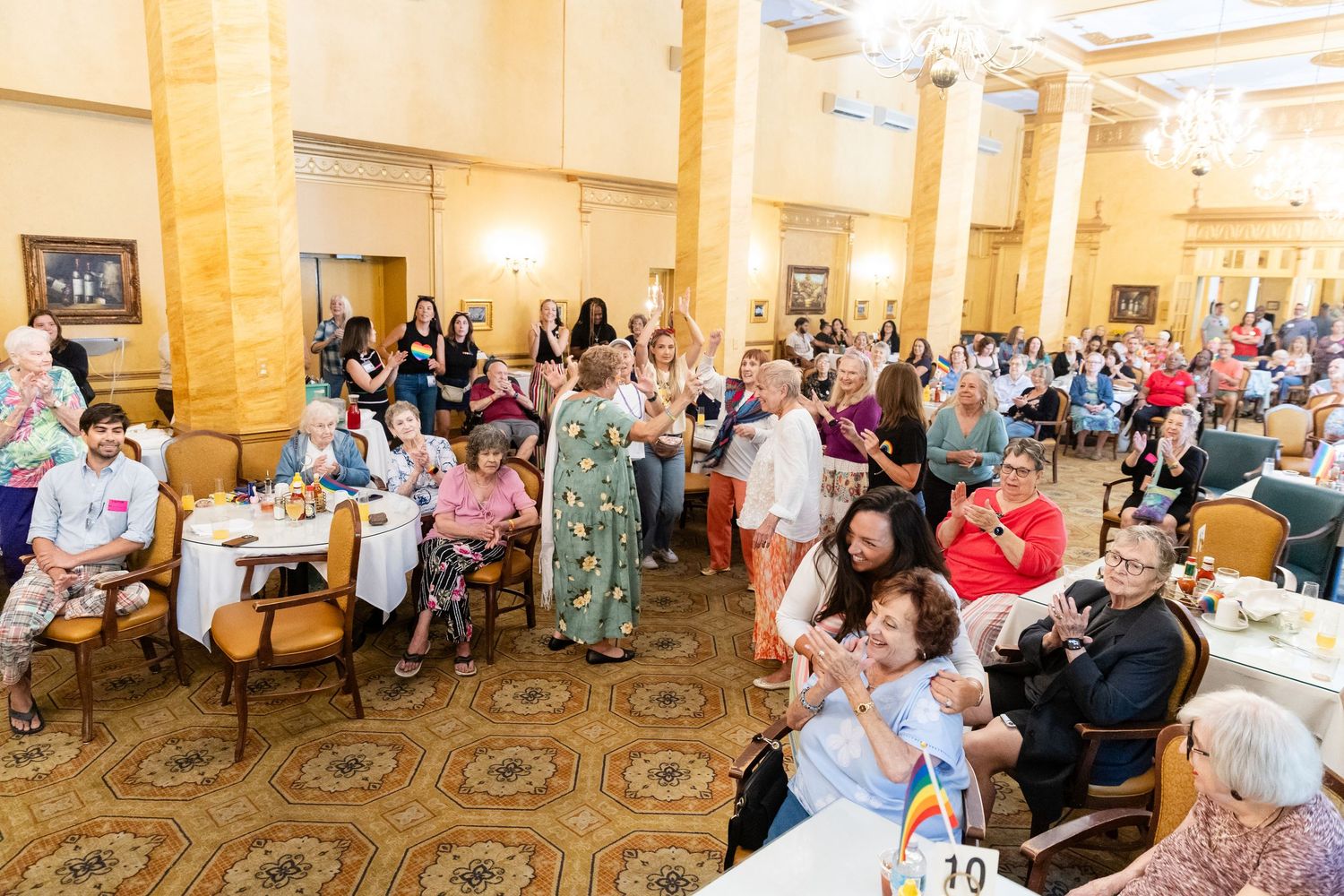Large crowd of seniors enjoy entertainment at a colorful Pride celebration in a ballroom.