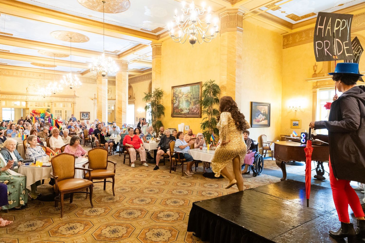 Drag performer entertains a crowd in an elegant ballroom decorated with a Happy Pride banner during a Pride celebration event.