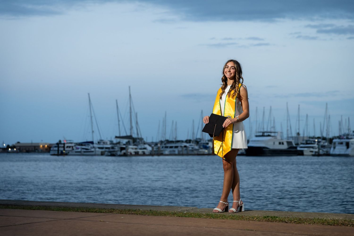 Young woman in white dress holding yellow graduation gown poses by marina at dusk with sailboats in background.