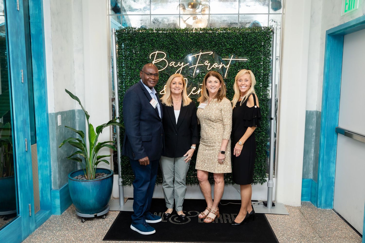 Four professionals posing together in front of a Bay Front venue green wall backdrop at a formal event.