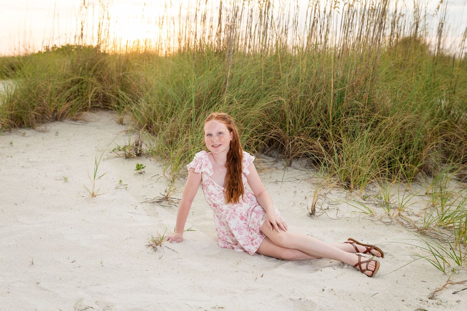 A smiling redhead girl in a floral dress sits on white sand dunes with sea oats at sunset.