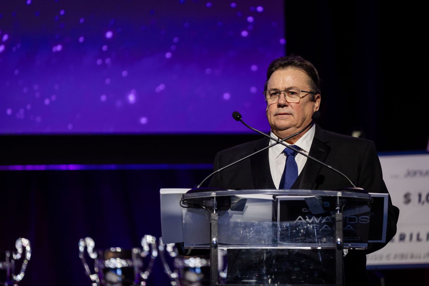 Man in black suit speaking at podium during formal awards ceremony with trophies displayed.