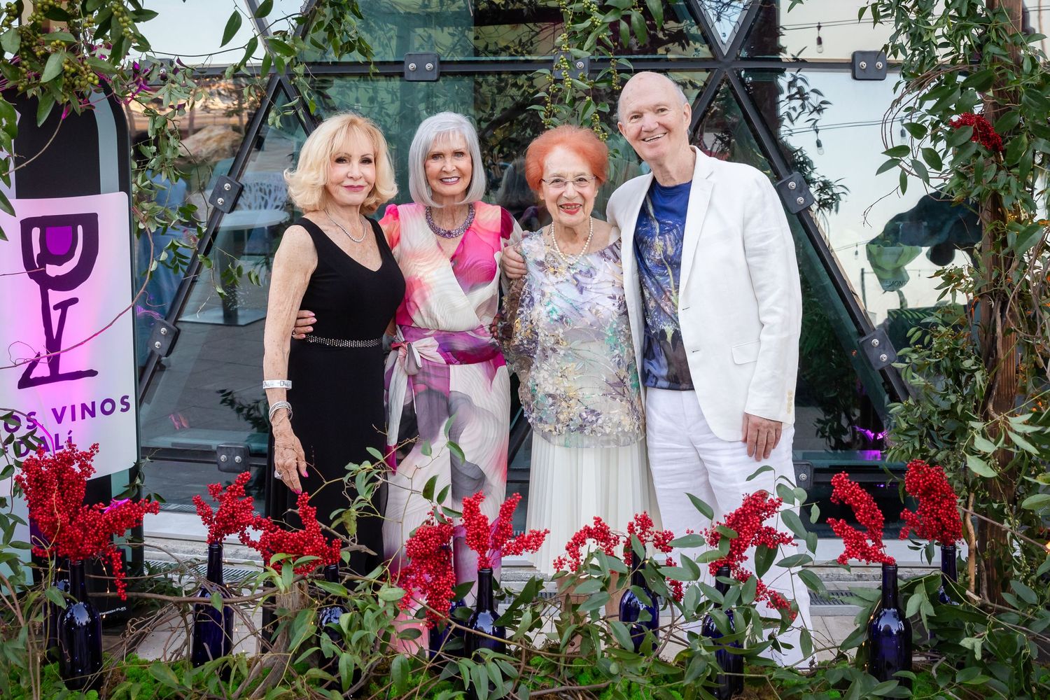 Four elegantly dressed women posing together at a wine event with floral decorations and wine bottles in background.