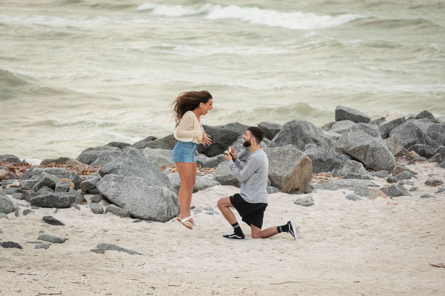 Man proposes to woman on sandy beach with rocky shoreline and ocean waves in the background.