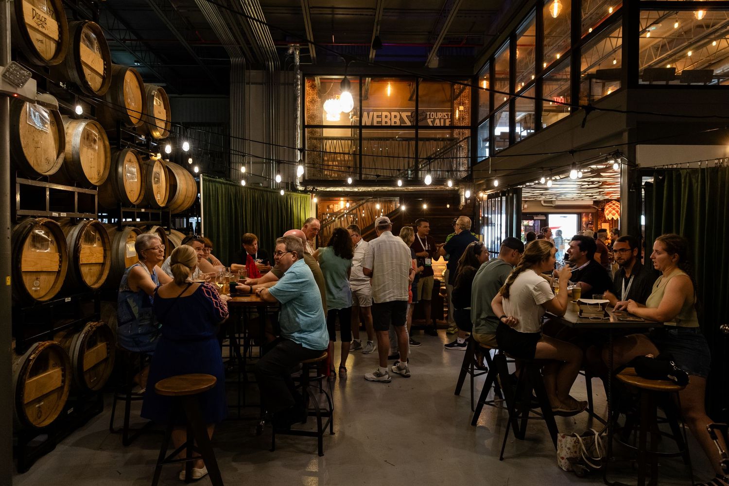 Busy craft brewery taproom with wooden barrels, string lights, and guests socializing at high-top tables.