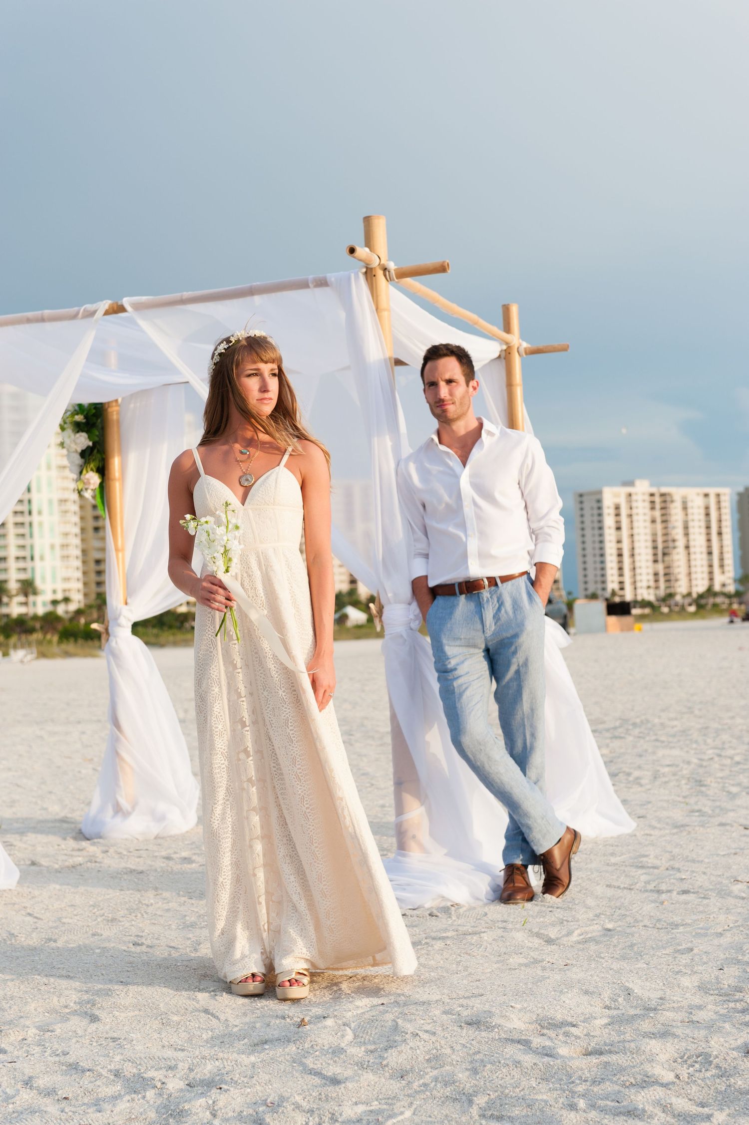 Bride and groom pose on sandy beach wedding venue with white draped bamboo arch and ocean backdrop.