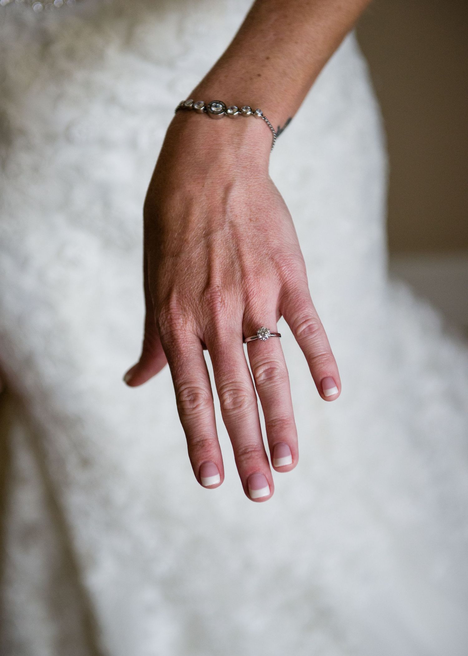 Bride showcasing diamond engagement ring and silver bracelet against white lace wedding dress.