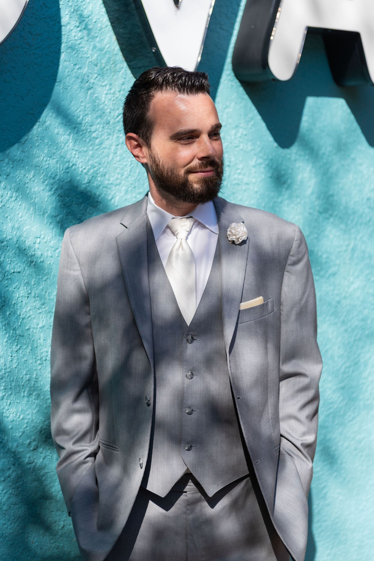 Groom in a gray three-piece suit with white tie and boutonniere posing against a teal wall.