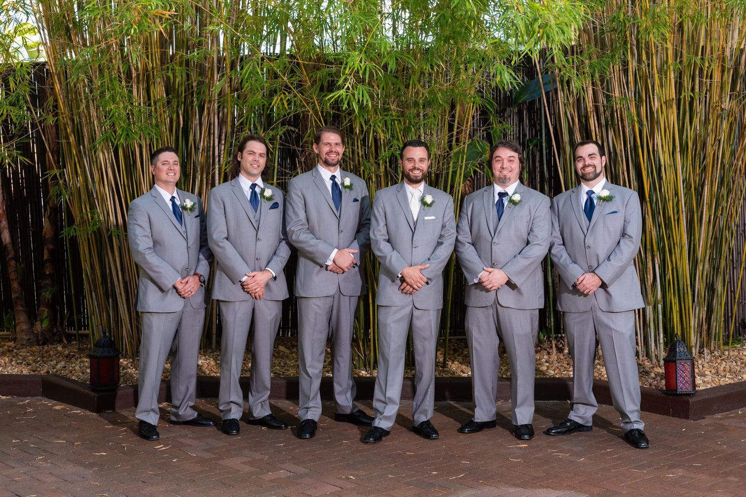 Six groomsmen in matching gray suits with boutonnieres pose outdoors in front of lush green bamboo at a wedding.