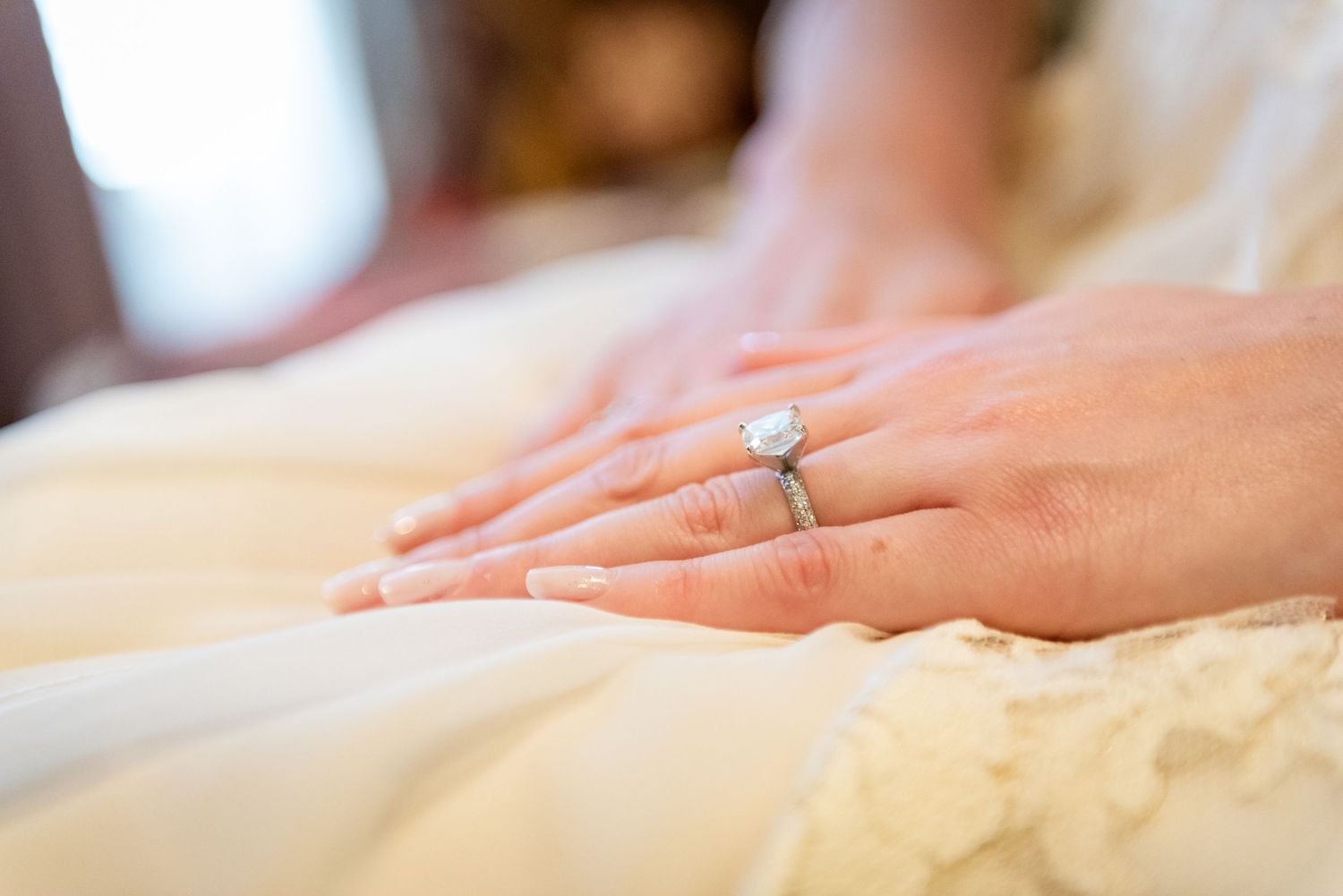 Close-up of bride's hands wearing a diamond engagement ring and wedding band on white dress.