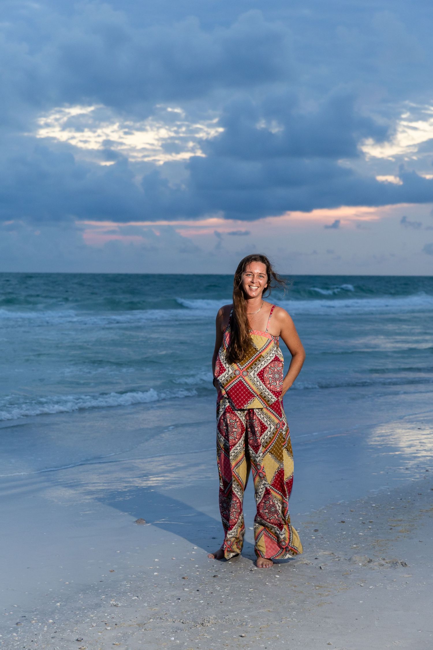 Young woman wearing vibrant patterned outfit posing on beach shoreline during blue hour with ocean waves.