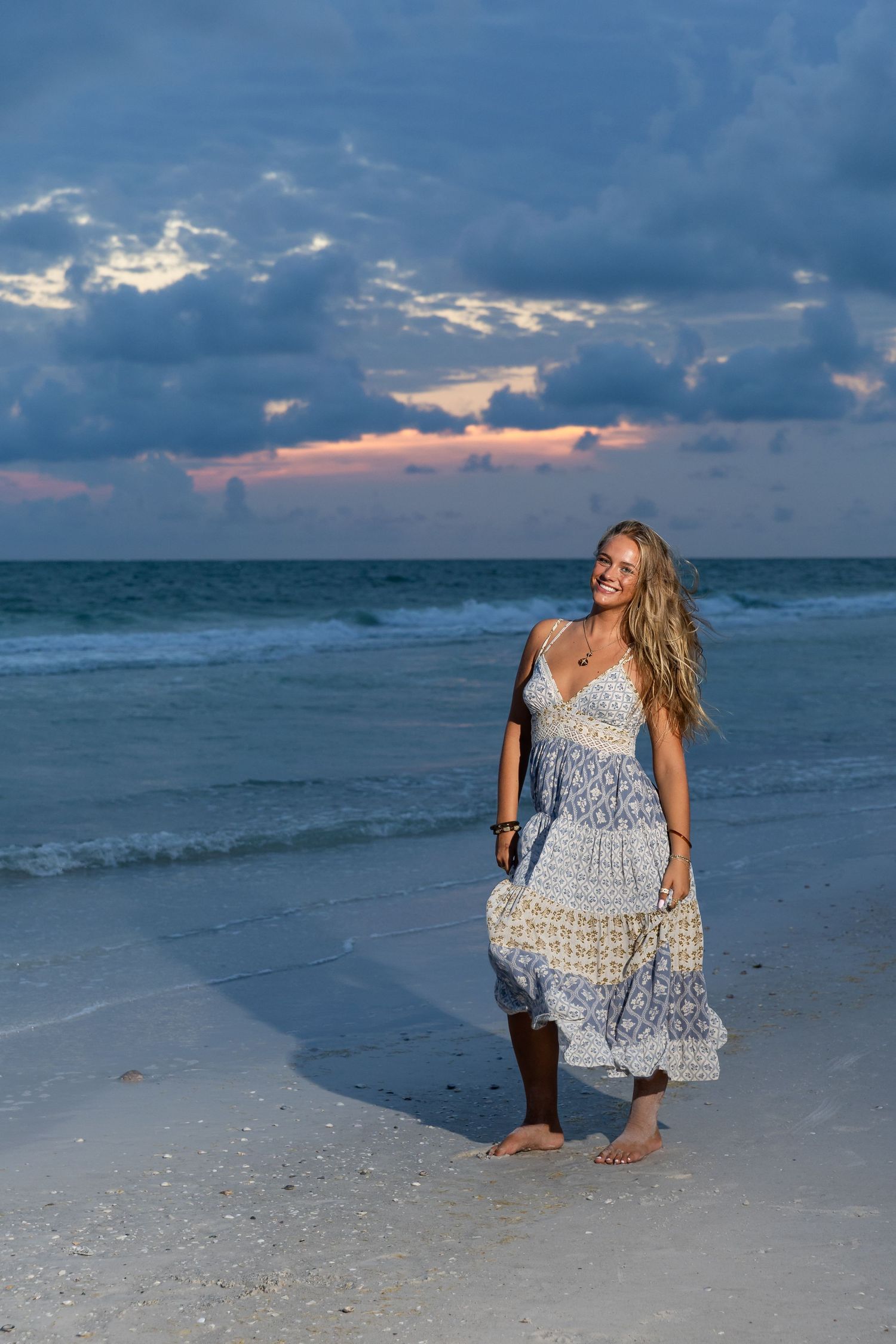 Smiling woman in boho maxi dress walking barefoot on sandy beach at sunset with dramatic cloudy sky.