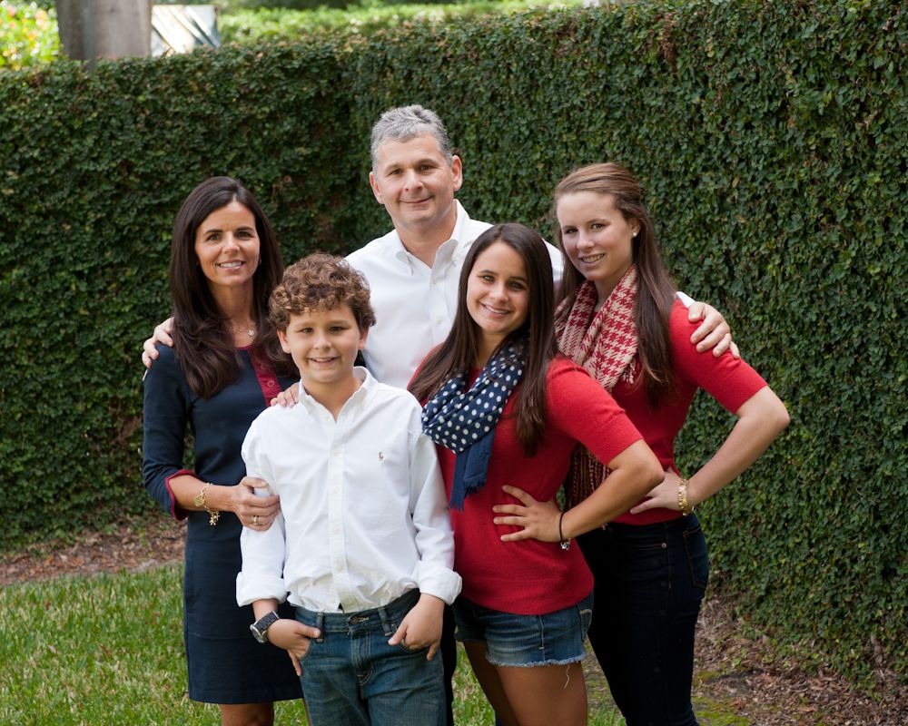Family of five posing outdoors in front of green hedge, smiling together in casual attire.