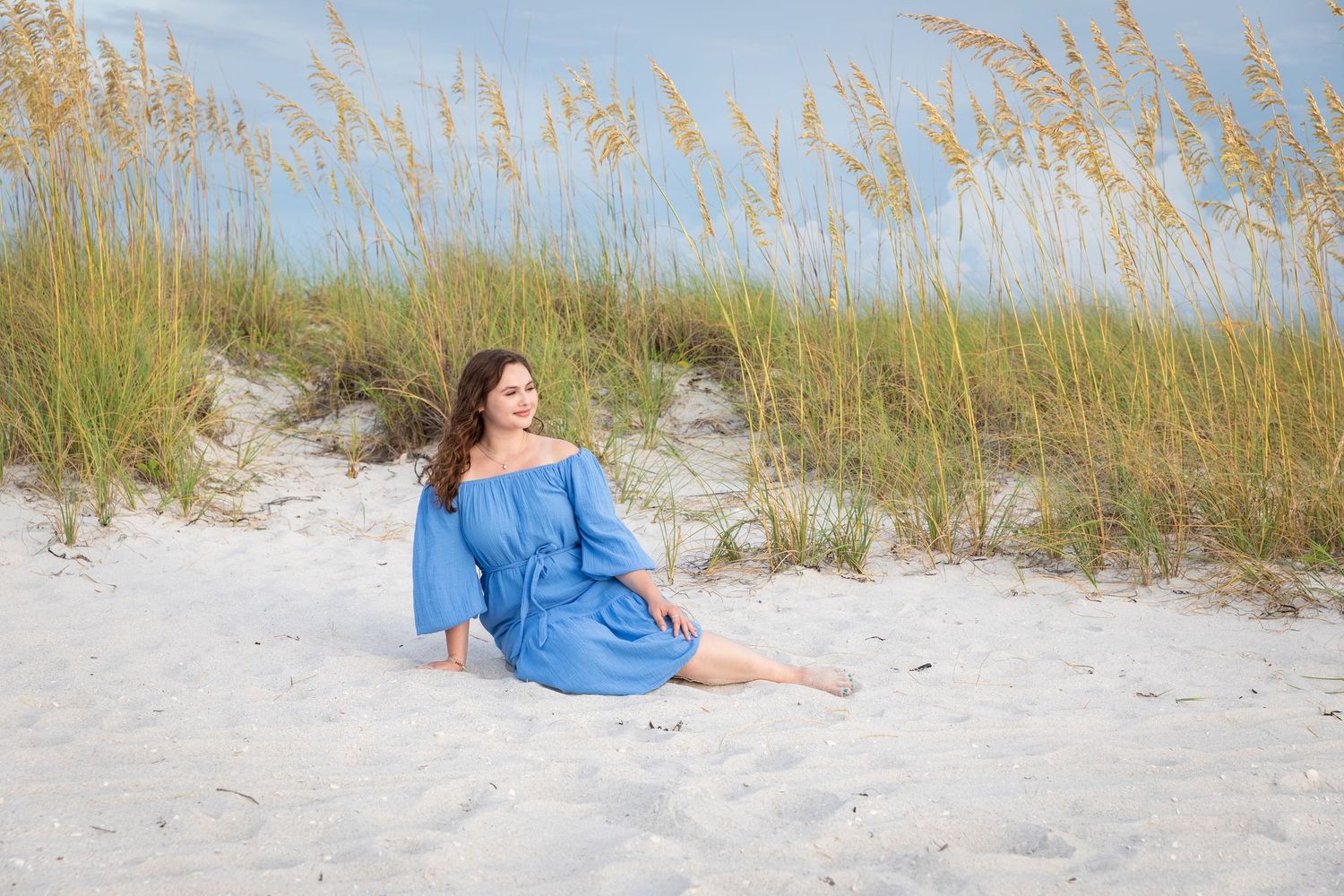 Young woman in blue off-shoulder dress sitting on white sand beach dunes with sea oats grass backdrop.