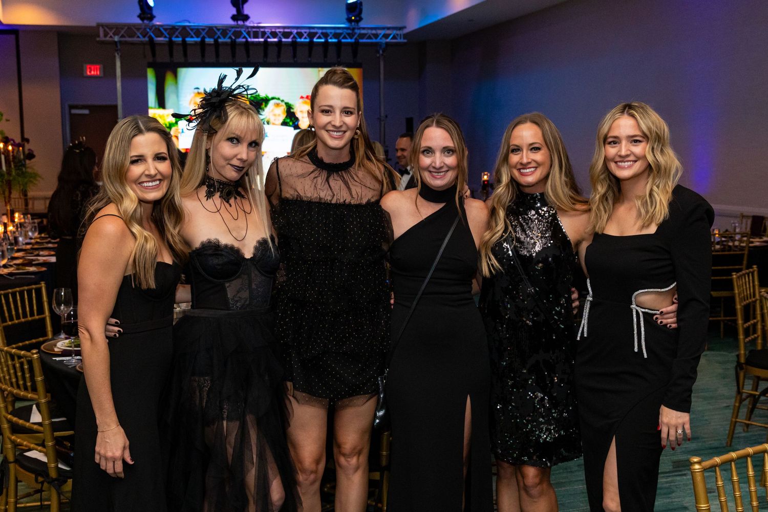 Six women in elegant black attire pose together at a formal gala event with gold chairs and decorative lighting.