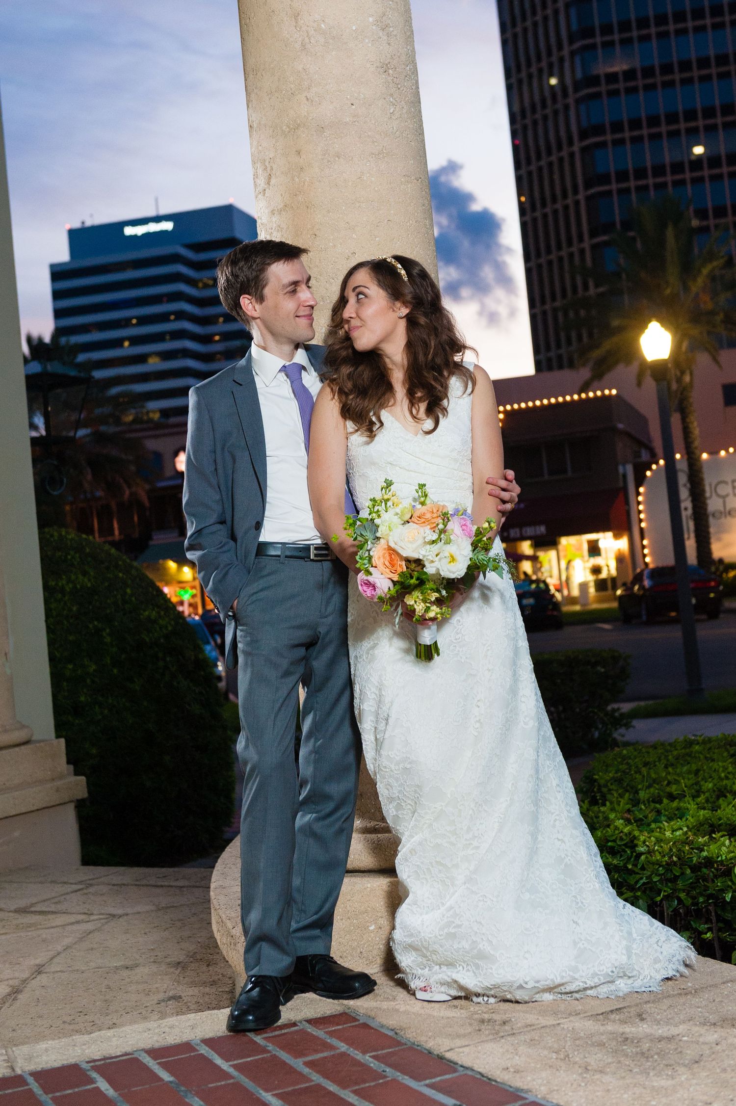 Bride and groom sharing romantic moment by column at dusk with city skyline in background.