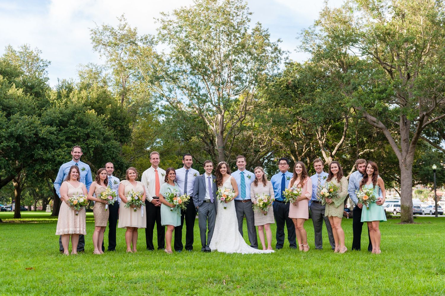 Large wedding party posing together on green lawn with trees in background on sunny day.
