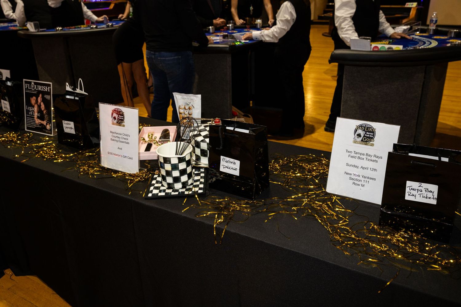 Black table with silent auction items, checkered boxes, and gold confetti at a casino-themed charity event.