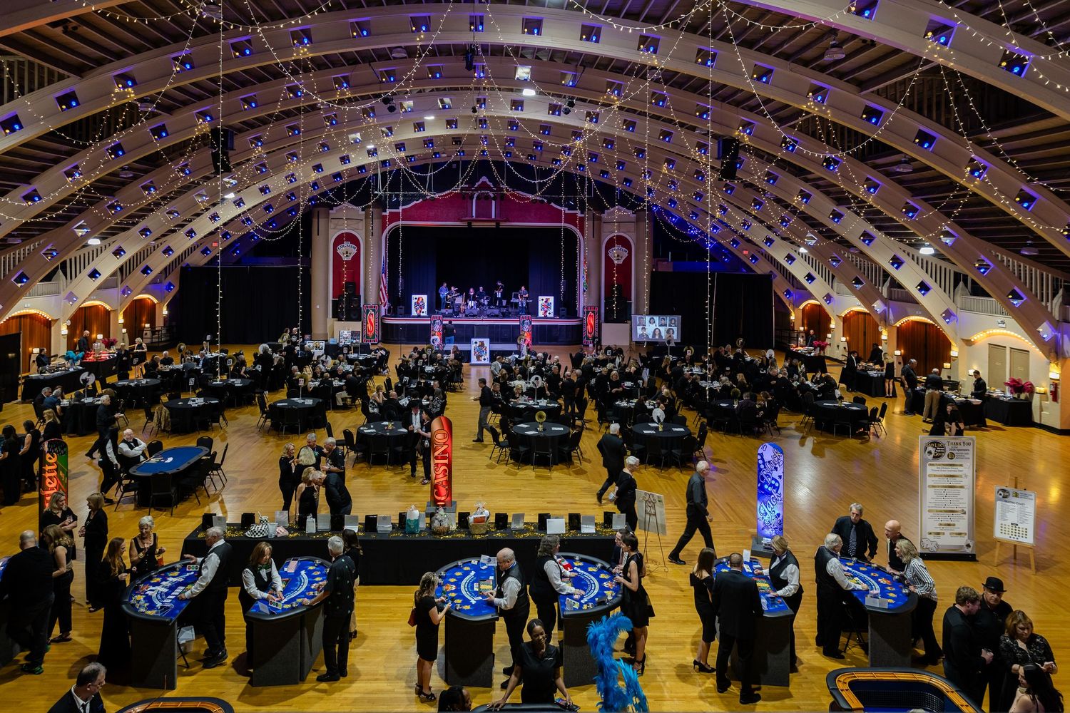 Elegant ballroom event with casino tables, guests mingling, and a stage with performers under an arched ceiling.