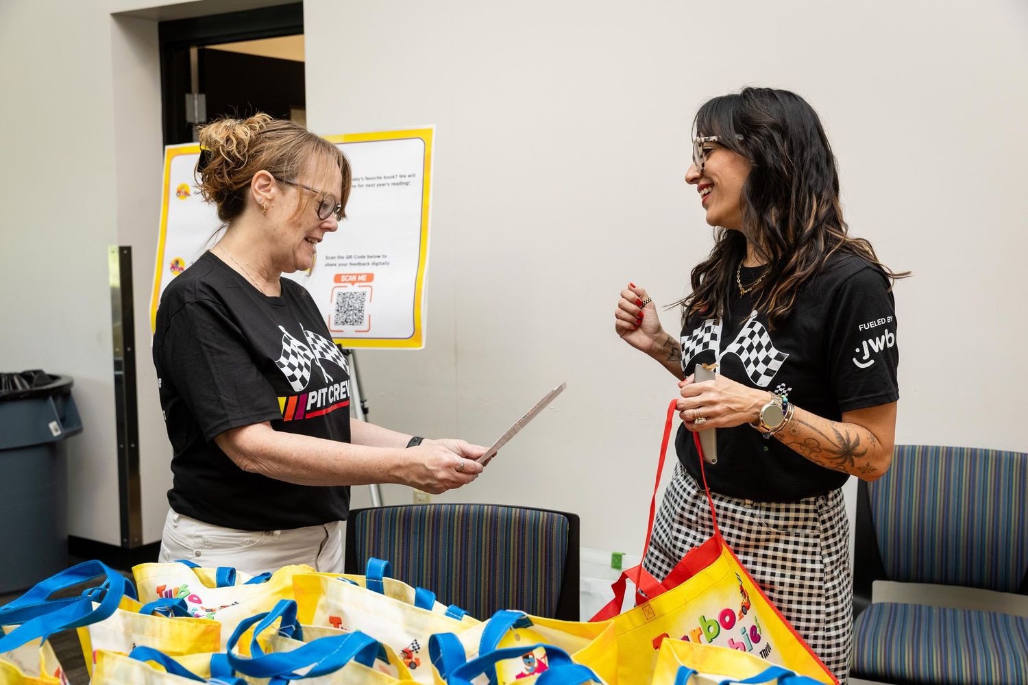 Two women in matching black t-shirts smile while reviewing materials at a table filled with colorful tote bags.