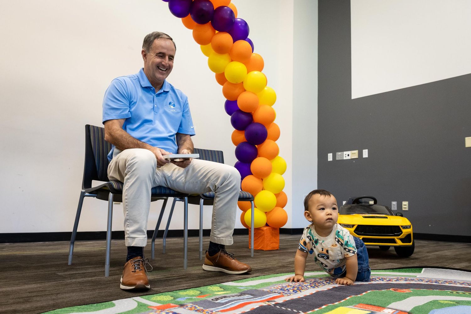 Man in blue shirt sits beside a baby playing on a colorful mat near an orange and purple balloon arch.