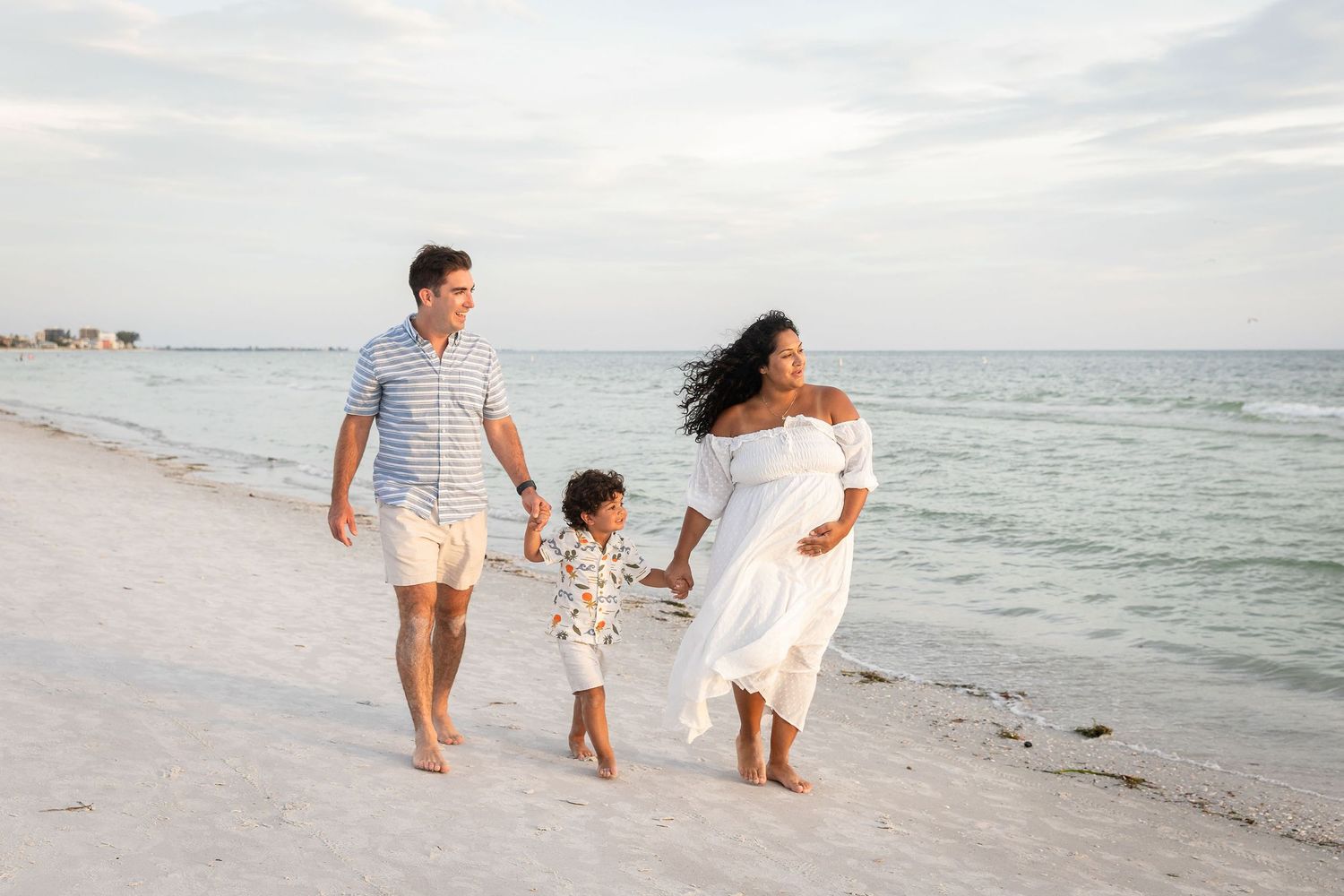 Pregnant woman in white dress walks on beach with husband and toddler, holding hands by calm ocean waters.