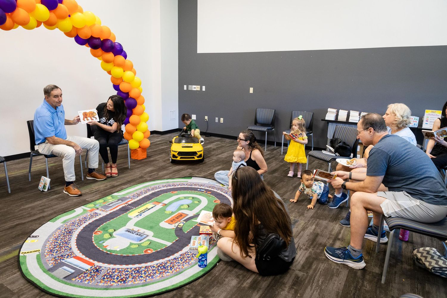 Children and families gather in a colorful room with balloon arch, race track mat, and yellow toy car for a kids event.